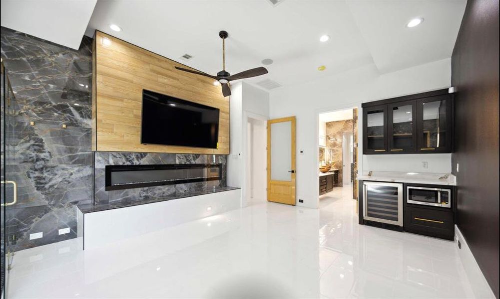 This modern master suite features glossy white floors, dark marble walls, and a wooden accent wall with a mounted TV above a sleek fireplace. It's equipped with high-end appliances including a mini fridge and microwave, complemented by dark cabinetry with gold accents. Bright lighting and a contrasting warm-toned door add to the contemporary aesthetic.