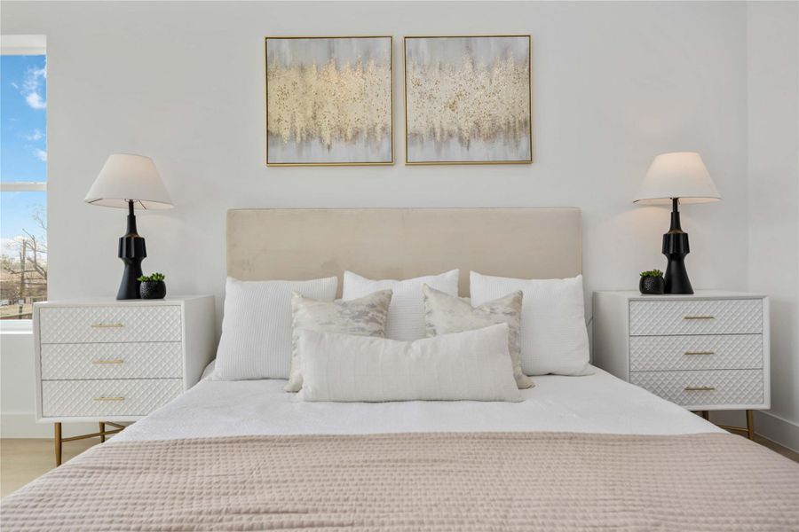 This elegantly staged primary suite features a plush upholstered headboard framed by matching modern nightstands, soft layered bedding, and contemporary artwork, creating a serene and sophisticated focal point within the space.