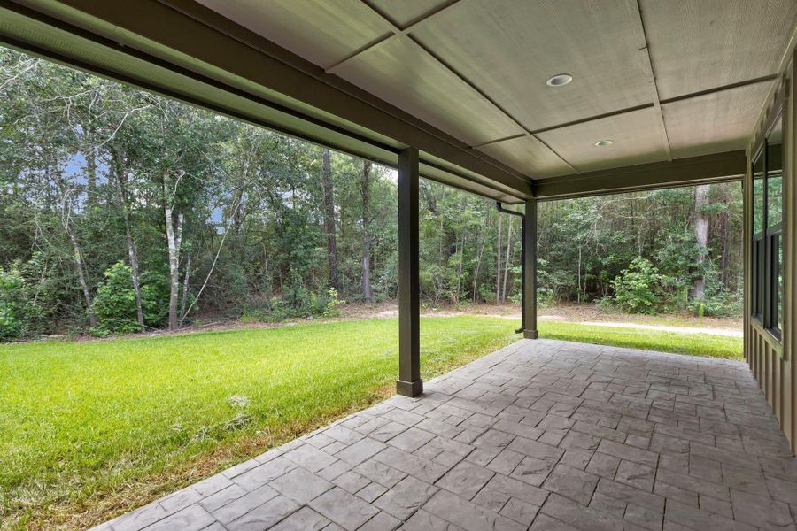 This photo showcases the 2nd covered patio with a stamped concrete floor and a wood-paneled ceiling, offering a view of a lush, green backyard bordered by trees, ideal for outdoor relaxation.