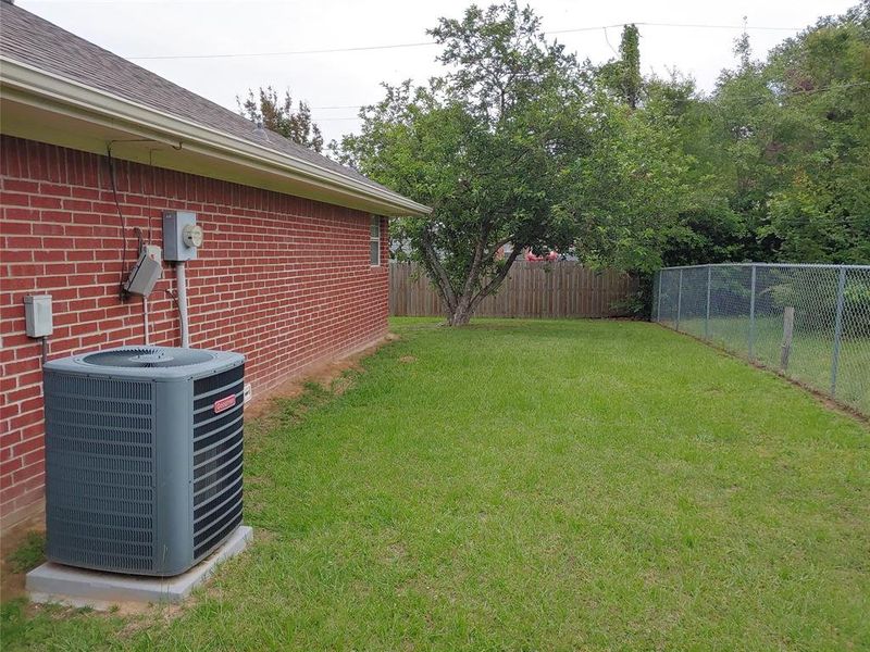 Expansive lawn area featuring a mature tree, a full chain-link fence, and a brick exterior building with a visible HVAC unit