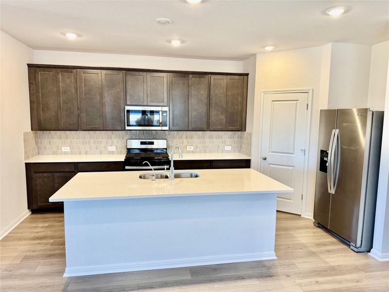 Kitchen featuring tasteful backsplash, stainless steel appliances, dark brown cabinets, light stone counters, and a kitchen island with sink Kitchen featuring tasteful backsplash, stainless steel appliances, dark brown cabinets, light stone counters, and a kitchen island with sink