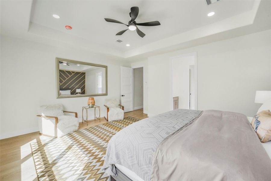 This spacious bedroom features a modern design with a large mirror, cozy seating area, and ample natural light. The room includes a ceiling fan, neutral tones, and a geometric-patterned rug, creating a comfortable and stylish atmosphere.