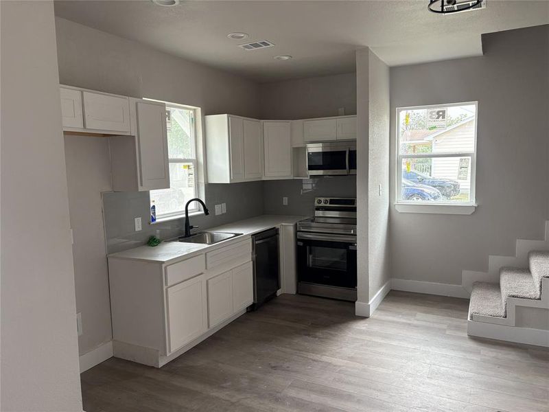 Modern kitchen featuring white cabinetry, light gray countertops, stainless steel appliances, a black gooseneck faucet, and wood-finish flooring Modern kitchen featuring white cabinetry, light gray countertops, stainless steel appliances, a black gooseneck faucet, and wood-finish flooring