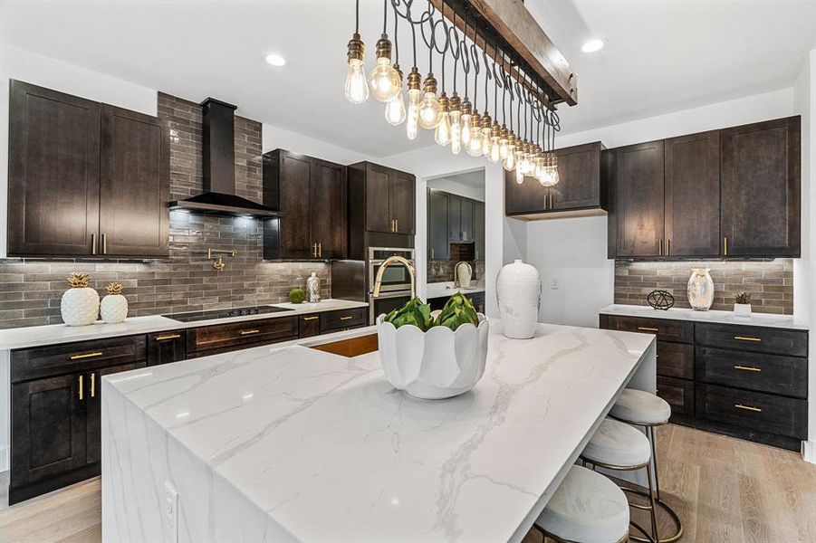 Kitchen with wall chimney range hood, hanging light fixtures, black electric stovetop, a center island with sink, and tasteful backsplash