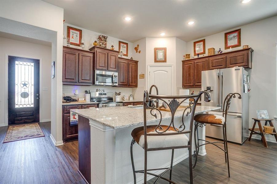 Kitchen with stainless steel appliances, a kitchen island with sink, a breakfast bar, tasteful backsplash, and dark wood-style floors Kitchen with stainless steel appliances, a kitchen island with sink, a breakfast bar, tasteful backsplash, and dark wood-style floors