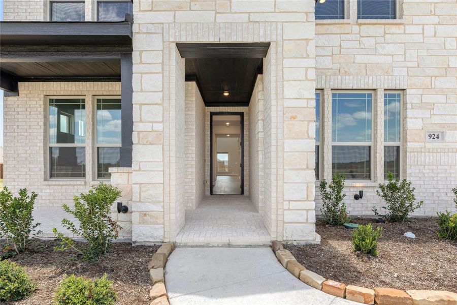 Front entry of a home with light stone exterior, covered walkway, and dark front door with landscaping Front entry of a home with light stone exterior, covered walkway, and dark front door with landscaping
