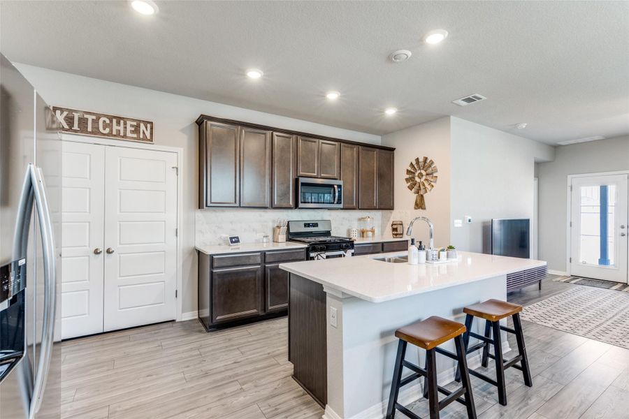 Kitchen featuring dark brown cabinets, backsplash, appliances with stainless steel finishes, a kitchen breakfast bar, and a kitchen island with sink