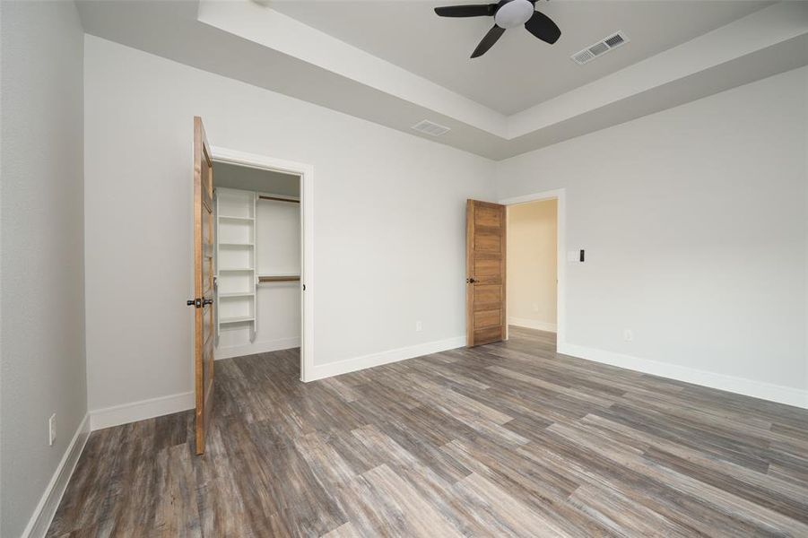 Unfurnished bedroom featuring a raised ceiling, a walk in closet, wood finished floors, and a ceiling fan