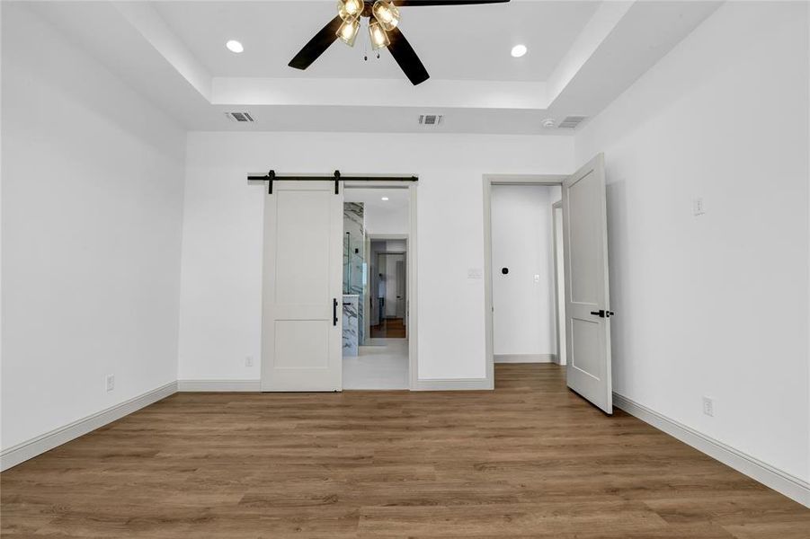 Unfurnished bedroom with a tray ceiling, ceiling fan, hardwood / wood-style flooring, and a barn door