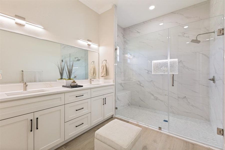 Spacious primary bathroom featuring a dual vanity with white cabinetry and quartz countertops, an expansive mirror, and a glass-enclosed shower with large format marble-look tile walls and a hexagonal tile floor
