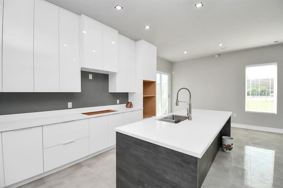 This is a modern, spacious kitchen featuring sleek white countertops, high-gloss cabinetry, and a large island with a built-in sink. The room is well-lit with natural light from the windows and recessed ceiling lights.