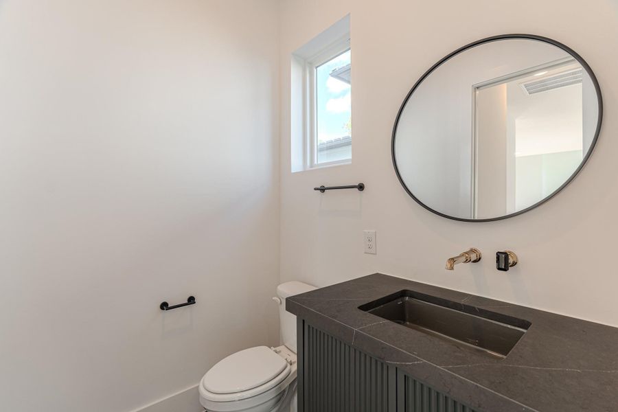 This bathroom features a sleek, modern design with a round mirror, wall-mounted faucet, and a stylish black countertop. It includes a toilet and a small window for natural light, enhancing its bright, minimalist aesthetic.