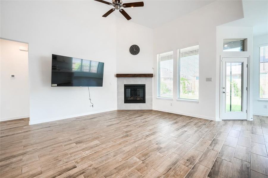 Unfurnished living room with a tiled fireplace, light wood-type flooring, a ceiling fan, and a high ceiling Unfurnished living room with a tiled fireplace, light wood-type flooring, a ceiling fan, and a high ceiling