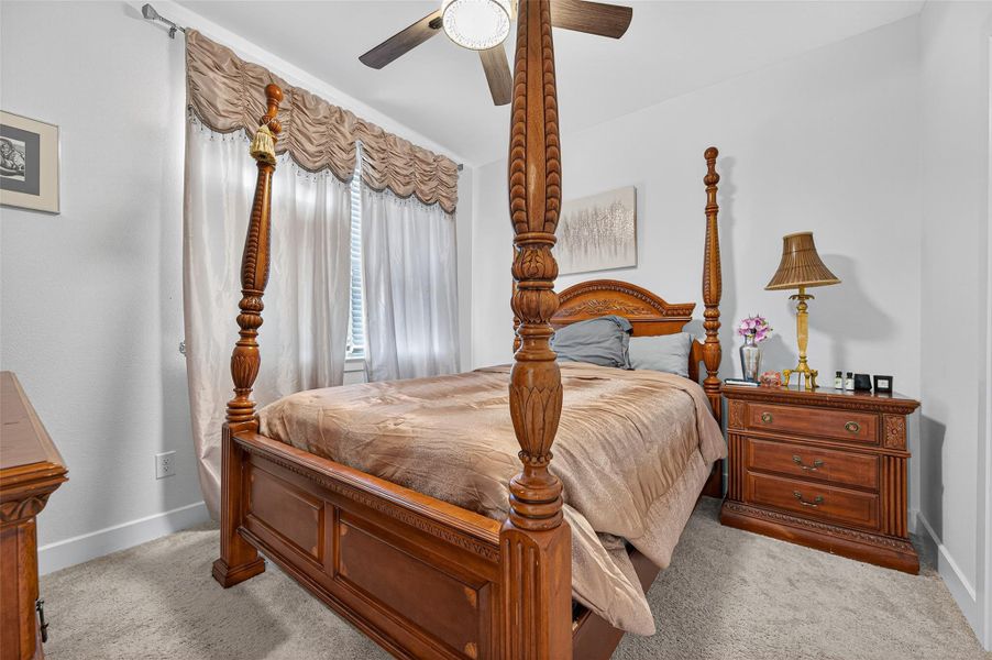 This cozy bedroom features a classic four-poster bed with elegant wood detailing, a ceiling fan, and soft carpeting. It includes a matching wooden nightstand with a decorative lamp and a large window adorned with stylish drapery, allowing for ample natural light. This cozy bedroom features a classic four-poster bed with elegant wood detailing, a ceiling fan, and soft carpeting. It includes a matching wooden nightstand with a decorative lamp and a large window adorned with stylish drapery, allowing for ample natural light.