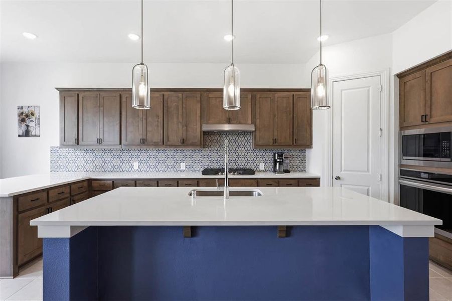 Kitchen featuring a large island with a white countertop and blue base, stainless steel appliances, wood cabinetry, and a patterned tile backsplash