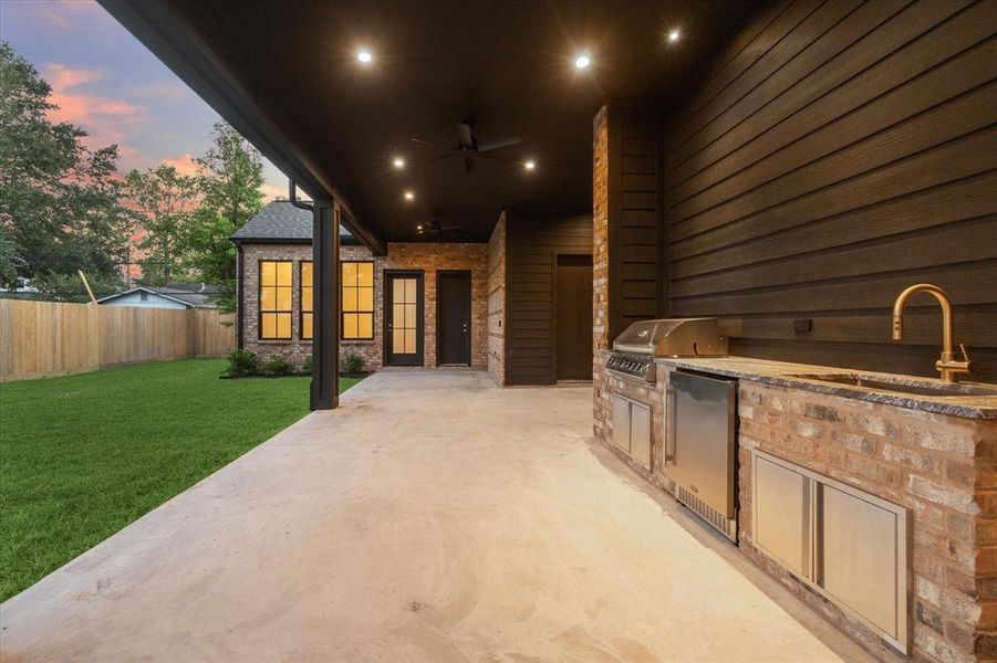 This outdoor covered patio is designed for both relaxation and entertaining. It features a full outdoor kitchen with built-in stainless steel appliances, including a 32" grill, outdoor fridge, and a sleek sink, all set against a backdrop of rustic brick and dark paneling. The patio extends out to a spacious lawn, fully fenced for added privacy. Recessed lighting and ceiling fans ensure comfort at any time of day or night, making this space perfect for gatherings or quiet evenings.