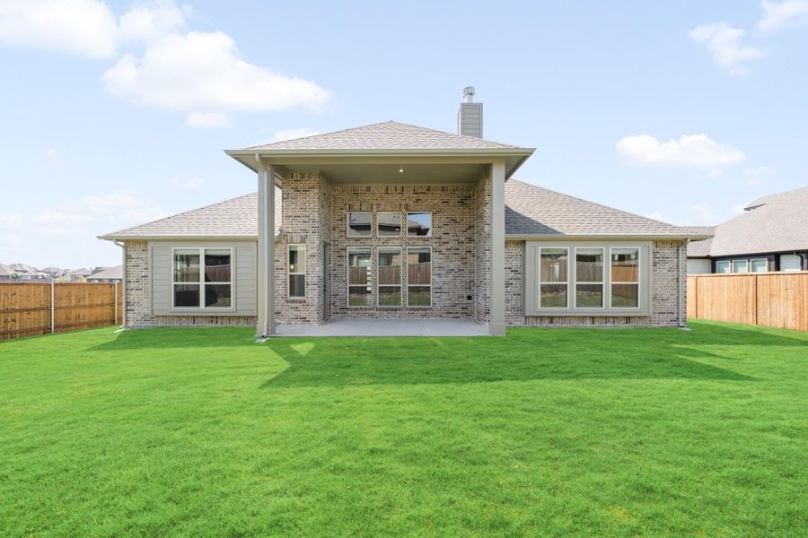 Rear view of brick home with covered patio, large windows, and fenced backyard with green lawn Rear view of brick home with covered patio, large windows, and fenced backyard with green lawn