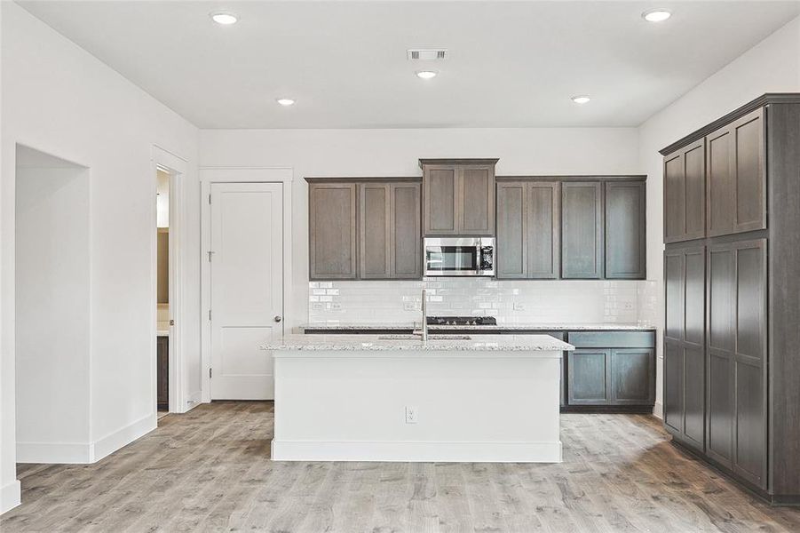 Kitchen featuring light wood-type flooring, a center island with sink, backsplash, and light stone counters