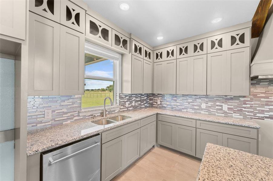 Kitchen featuring stainless steel dishwasher, custom exhaust hood, tasteful backsplash, sink, and light stone countertops