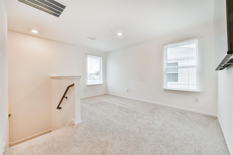 Bright and spacious carpeted room with two windows, offering ample natural light. Features a stairway with a wooden handrail and recessed lighting for a cozy ambiance. Perfect for a versatile living area.