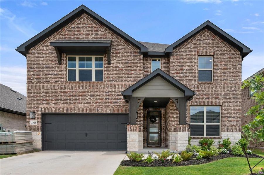 Two-story brick facade featuring a dark gray garage door, covered entry with stone accents, dark trim, and an awning window feature