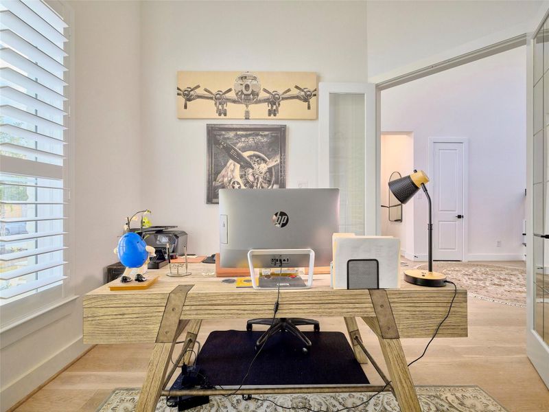 Modern home office featuring expansive windows, elegant shutters, wide plank flooring, and custom built-in shelving for a stylish and functional workspace.