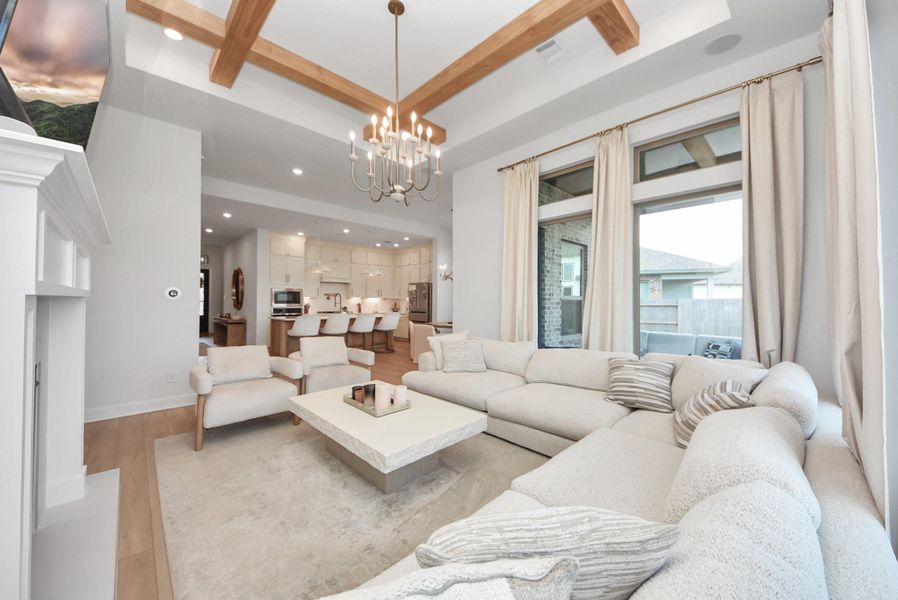 Spacious and inviting living area with elegant wood beam ceilings, a cozy sectional, and a modern chandelier, seamlessly flowing into a bright, open kitchen.