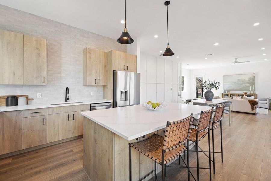 Kitchen featuring light brown cabinetry, a kitchen breakfast bar, decorative light fixtures, stainless steel appliances, and light stone counters featuring modern glass door, dusk to dawn motion detection sconce, and ring doorbell, and board and batten detail