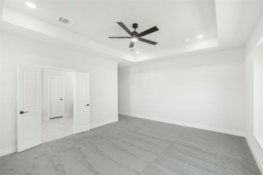 Empty bedroom with tray ceiling, ceiling fan, gray carpet, and two white doors leading to bathroom and closet Empty bedroom with tray ceiling, ceiling fan, gray carpet, and two white doors leading to bathroom and closet