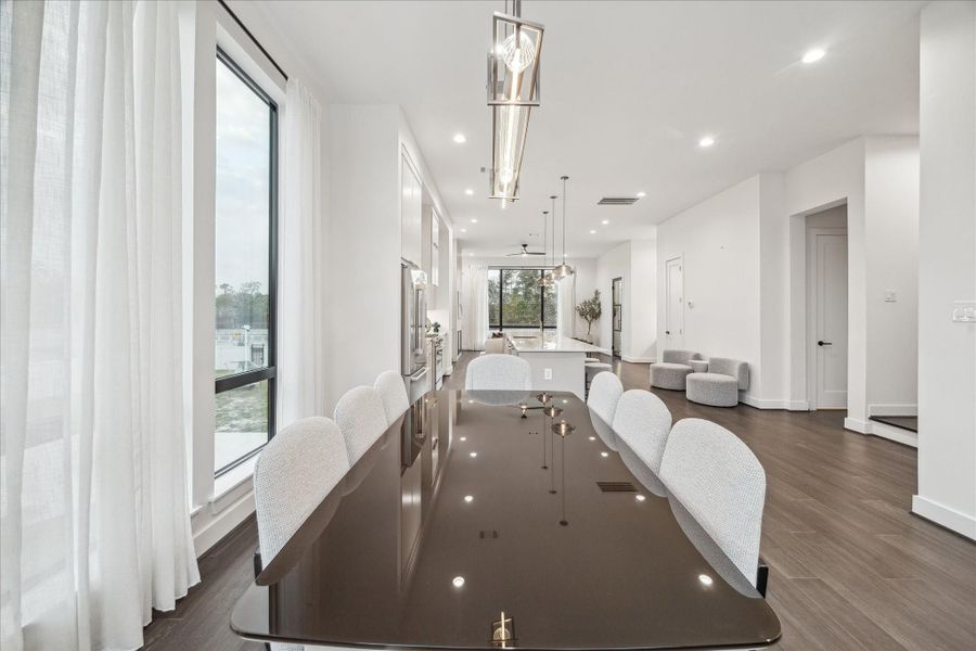 Expansive open-concept layout framed by soaring ceilings and wide-plank flooring, seamlessly connecting dining, kitchen, and living areas. Oversized windows dressed in soft panels invite abundant natural light while showcasing treetop views. Linear architectural lighting enhances the modern aesthetic, and the elongated sightlines emphasize the depth and scale of this thoughtfully designed second-floor gathering space.