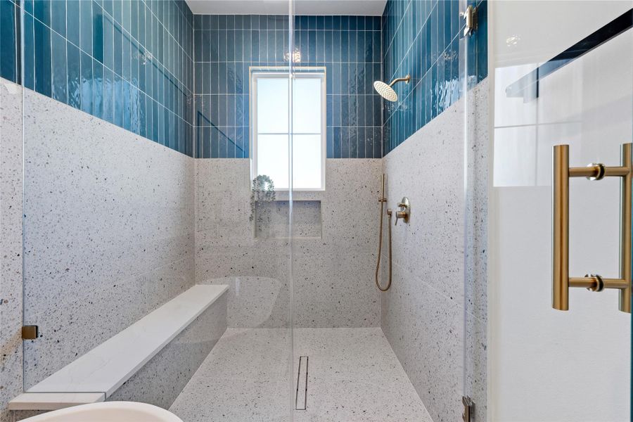 A frameless glass partition defines the spacious walk-in shower, which includes a built-in bench and linear floor drain. The architecture utilizes a central window to flood the tiled space with natural light.