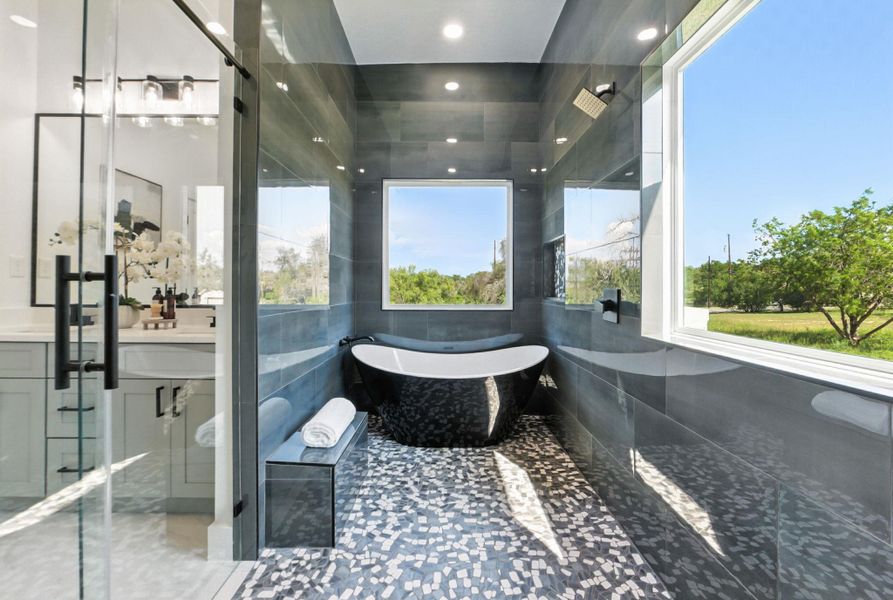 A modern spa-like bathroom featuring a sleek freestanding black bathtub, large windows with scenic views, glossy gray tile walls, and a mosaic tile floor.
