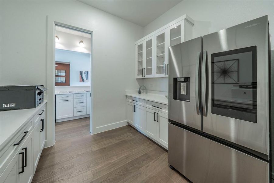 Kitchen featuring stainless steel refrigerator with ice dispenser, dark wood-style flooring, glass insert cabinets, white cabinetry, and light countertops Kitchen featuring stainless steel refrigerator with ice dispenser, dark wood-style flooring, glass insert cabinets, white cabinetry, and light countertops