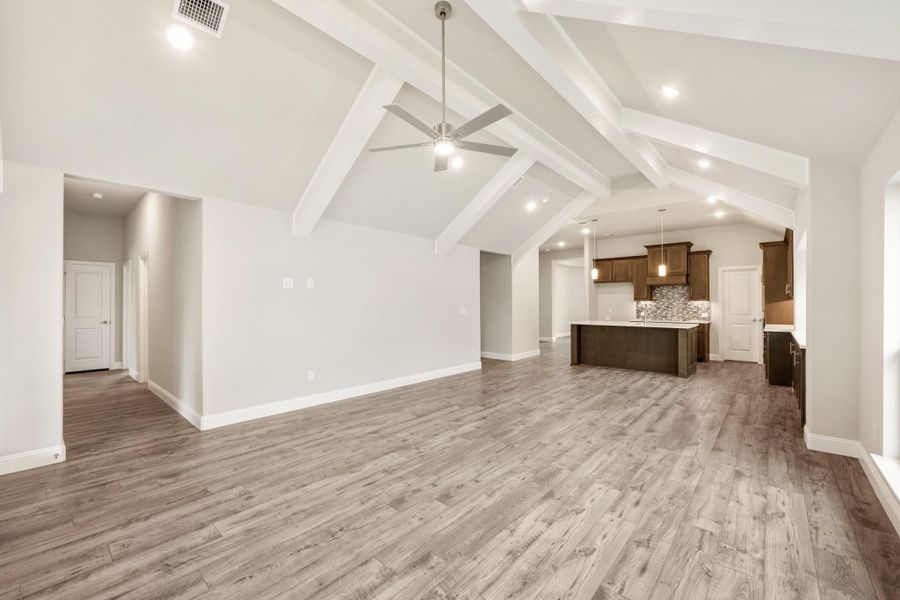 Open living area with vaulted beamed ceiling, ceiling fan, hardwood floors, and kitchen with dark cabinets in background