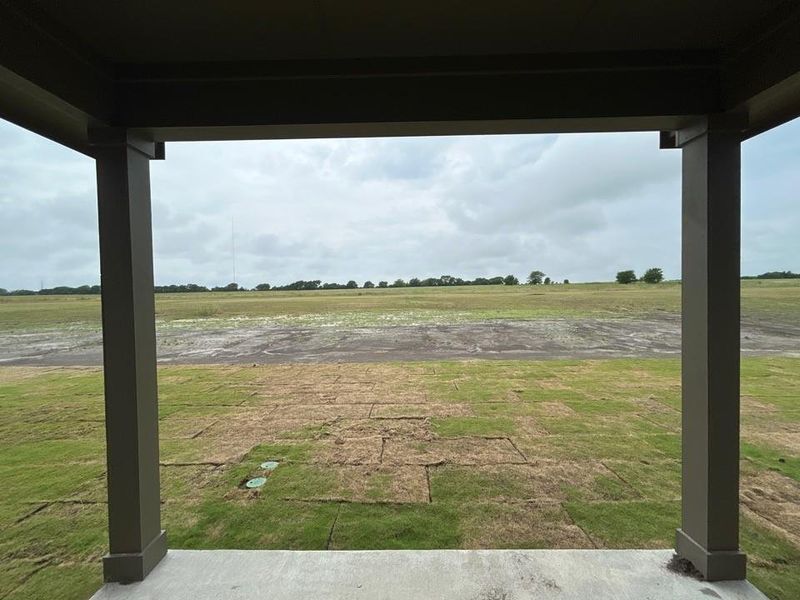 Covered patio with concrete flooring overlooking an expansive green space with partial sod installation and distant trees Covered patio with concrete flooring overlooking an expansive green space with partial sod installation and distant trees