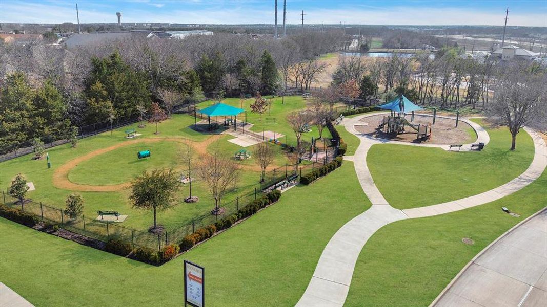 Expansive green space featuring a shaded playground, a fenced pet park with an agility tunnel, a covered picnic area, and paved walking paths