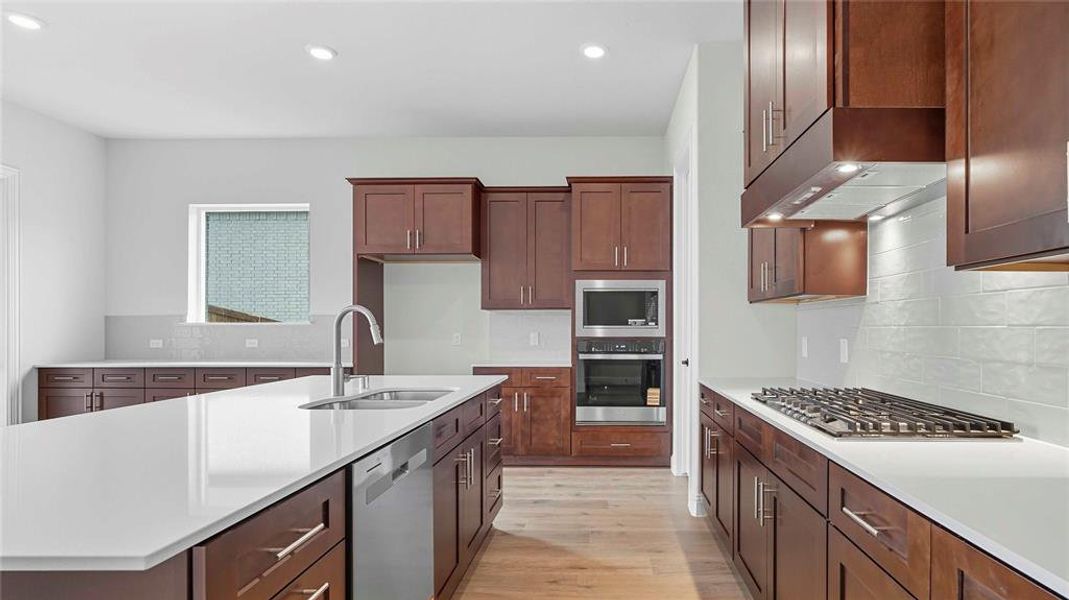 Kitchen featuring a large central island with an undermount sink and dishwasher, rich wood-finish cabinetry, light-toned countertops, and a white subway tile backsplash