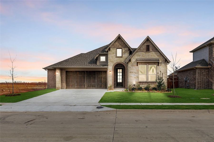 Single-story home with stone and brick exterior, two-car garage, arched entry, and green front lawn at dusk Single-story home with stone and brick exterior, two-car garage, arched entry, and green front lawn at dusk