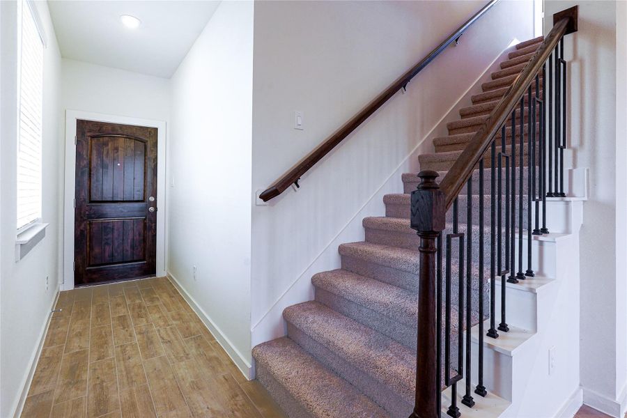 Bright entryway featuring a dark wooden front door, wood-tile flooring, and a carpeted staircase with elegant railing, leading to the upper floor.