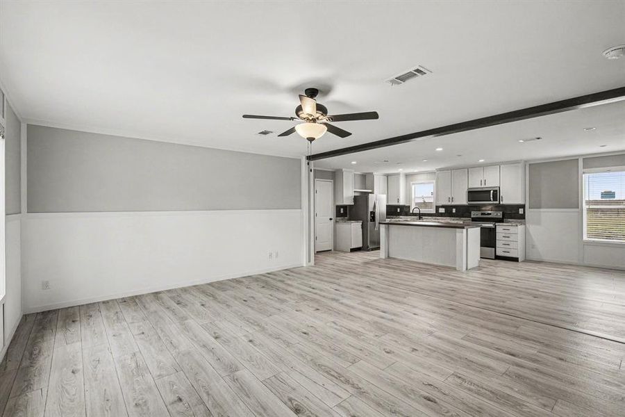 Spacious open-concept living area featuring wood-look flooring, a ceiling fan with integrated lighting, and a kitchen with white cabinetry, dark countertops, and stainless steel appliances