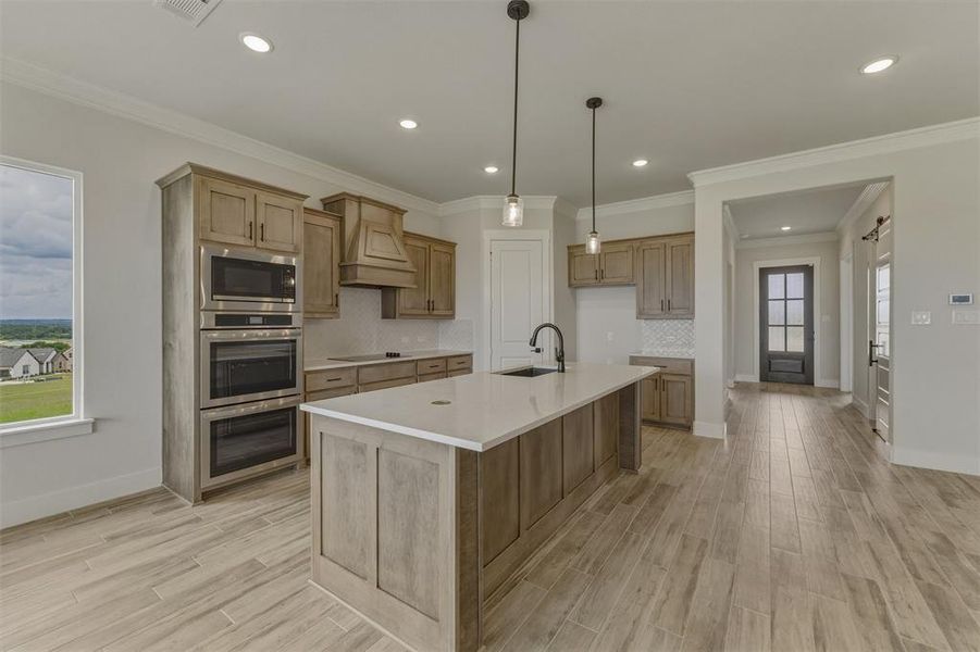 Spacious kitchen featuring a central island with sink, light wood-finish cabinetry, built-in stainless steel double ovens, and wood-finish flooring Spacious kitchen featuring a central island with sink, light wood-finish cabinetry, built-in stainless steel double ovens, and wood-finish flooring