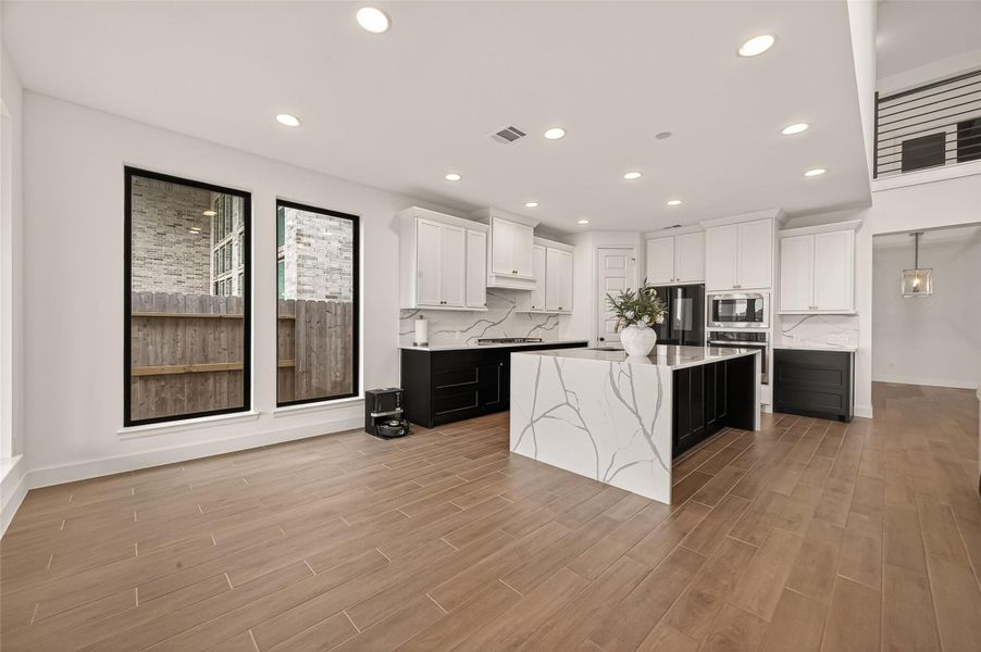 This modern kitchen features a spacious open layout with sleek white cabinetry, a large marble island, and stainless steel appliances. The space is brightened by large windows and recessed lighting, with wood-style flooring adding warmth.
