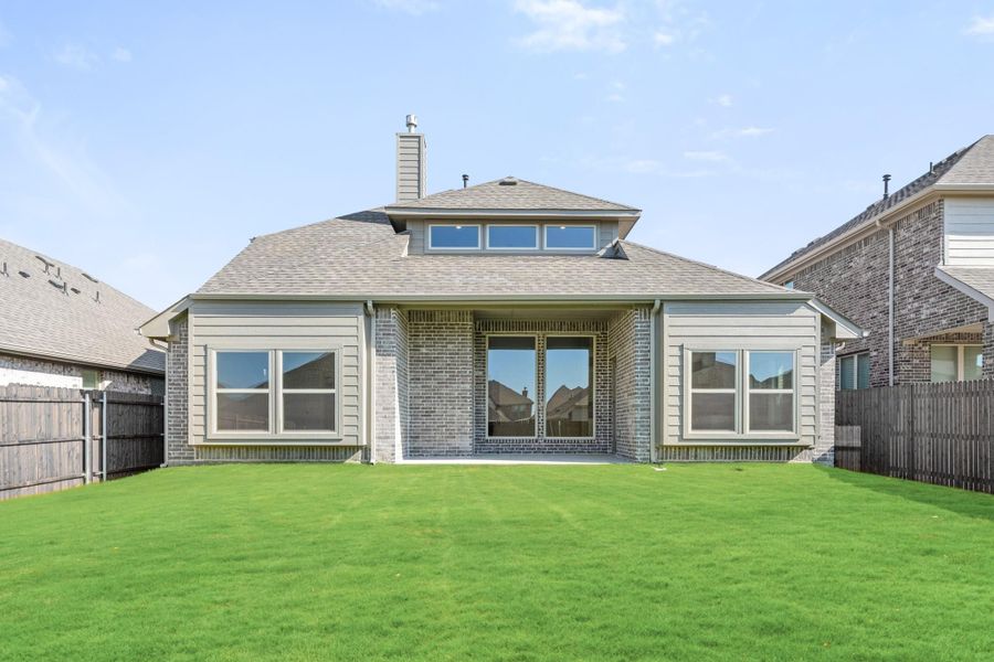 Rear exterior of single-story brick home with covered patio, large windows, and fenced backyard with green lawn. Rear exterior of single-story brick home with covered patio, large windows, and fenced backyard with green lawn.