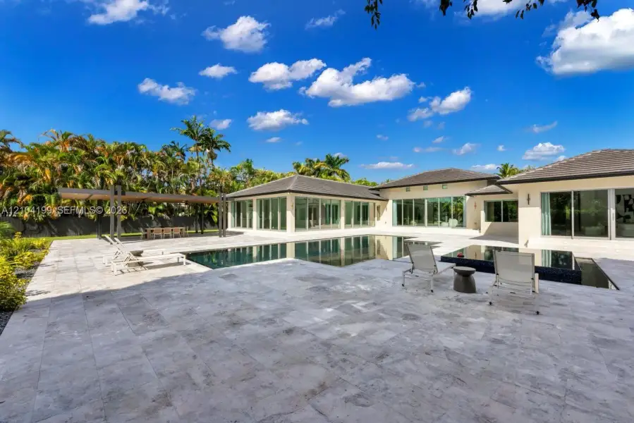 Expansive stone patio featuring a multi-level swimming pool with a raised spa, built-in covered pergola, and floor-to-ceiling glass wall panels