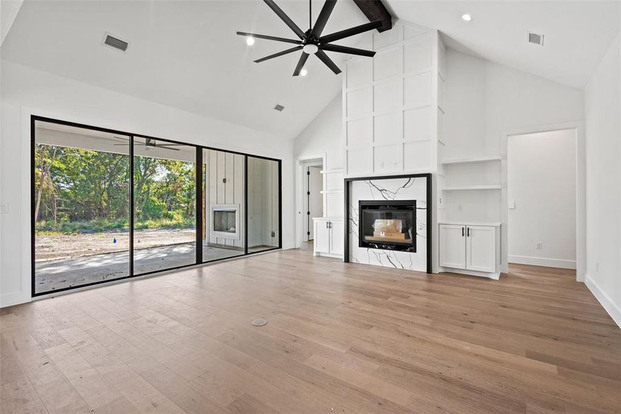 Unfurnished living room featuring a premium fireplace, high vaulted ceiling, beam ceiling, light wood-style flooring, and ceiling fan Unfurnished living room featuring a premium fireplace, high vaulted ceiling, beam ceiling, light wood-style flooring, and ceiling fan