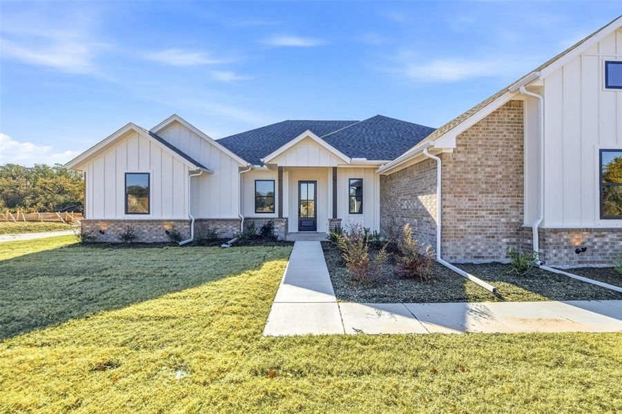 Modern farmhouse with board and batten siding, a front lawn, roof with shingles, and brick siding Modern farmhouse with board and batten siding, a front lawn, roof with shingles, and brick siding
