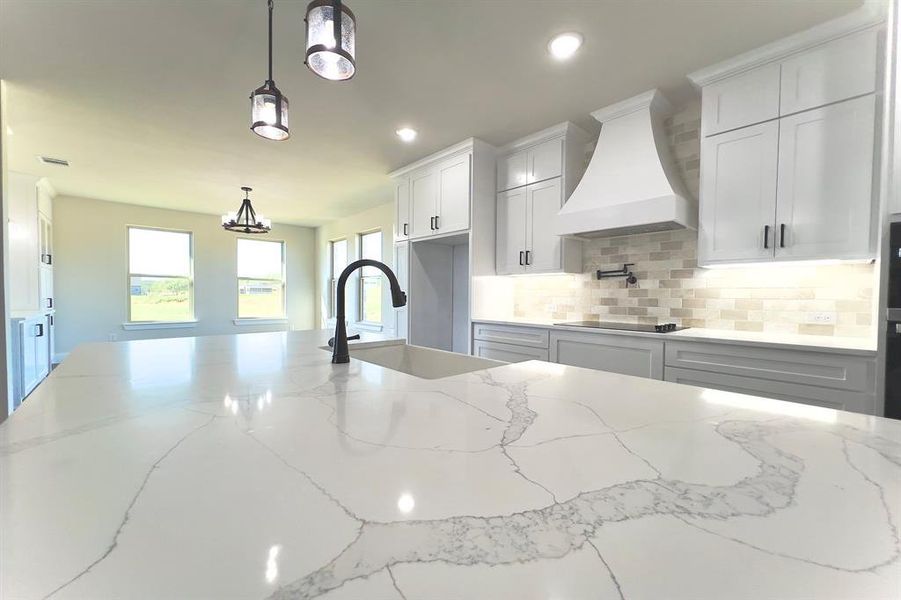 Kitchen featuring light stone countertops, tasteful backsplash, custom exhaust hood, hanging light fixtures, and white cabinetry