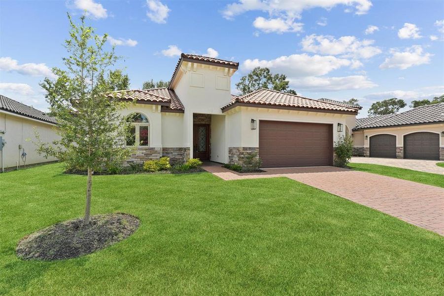 This is a single-story home with a Mediterranean-style tiled roof, featuring a well-manicured lawn, paver driveway, and an attached garage. The exterior has a mix of stucco and stone accents, giving it a modern yet classic look.