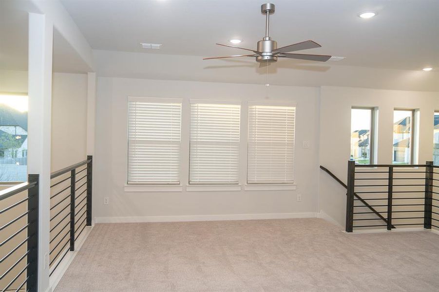Carpeted loft space featuring multiple windows, recessed lighting, a modern ceiling fan, and black metal railing Carpeted loft space featuring multiple windows, recessed lighting, a modern ceiling fan, and black metal railing