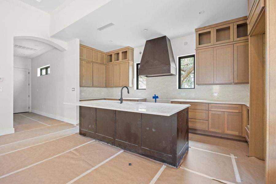 Kitchen featuring a large central island with a light-toned countertop, a custom range hood, shaker-style cabinetry with upper glass-front cabinets, and a contemporary sink faucet